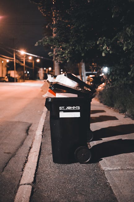 A black wheeled rubbish bin situated on the edge of a residential pavement during nighttime, with the label 'ST. JOHN'S' visible on the front. The bin's lid is open, revealing a mixed collection of waste and cardboard boxes inside, some leaning out slightly. Surrounding the bin, there are dark shadows cast by nearby trees and bushes on the pavement. In the background, warm street lighting illuminates the quiet street, with streetlights lining the road and a few parked cars visible further down. The scene depicts typical urban waste disposal, possibly used for private or alternative rubbish collection outside regular council services, consistent with the context of independent waste handling. The weather appears clear, and the environment is calm, emphasizing the bin's role in waste management on a residential street at night.