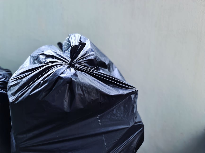 A black wheeled rubbish bin situated on the edge of a residential pavement during nighttime, with the label 'ST. JOHN'S' visible on the front. The bin's lid is open, revealing a mixed collection of waste and cardboard boxes inside, some leaning out slightly. Surrounding the bin, there are dark shadows cast by nearby trees and bushes on the pavement. In the background, warm street lighting illuminates the quiet street, with streetlights lining the road and a few parked cars visible further down. The scene depicts typical urban waste disposal, possibly used for private or alternative rubbish collection outside regular council services, consistent with the context of independent waste handling. The weather appears clear, and the environment is calm, emphasizing the bin's role in waste management on a residential street at night.