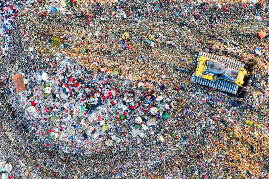 An aerial view of a large landfill site filled with a dense layer of mixed waste materials, including plastic, paper, and metal debris, with noticeable patches of colored plastics and foam. In the top right corner, a yellow tracked excavator is visible, partially buried in the waste, engaged in moving or managing the accumulated rubbish. The terrain is uneven and layered with various types of refuse, creating a textured surface. Surrounding debris shows signs of weathering, and the waste extends beyond the frame, indicating the scale of waste accumulation. The environment appears outdoor, with natural lighting highlighting the various colors and textures of the waste. This scene relates to waste management and rubbish disposal services, reflecting the volume of waste that independent collections, such as those offered by Waste Collection Ealing, aim to manage efficiently and responsibly within private or alternative waste handling contexts.