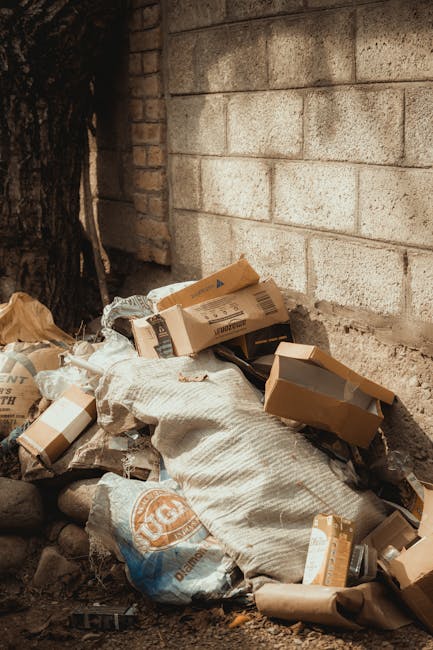 A small pile of mixed waste materials located outdoors alongside a brick and concrete wall. The heap includes flattened cardboard boxes, some with visible printed labels and logos, a large white cloth or sack partially covering the debris, and a plastic bag with a blue and orange logo. The waste is resting on uneven ground with small rocks and dirt, near the base of a tree trunk on the left side of the image. The brick wall behind the pile is weathered and unpainted, and the scene appears to be an outdoor area suitable for independent waste collection or private rubbish clearance services. The lighting is natural but subdued, providing clear visibility of textures and materials. This scene exemplifies a typical scene where a waste removal company, such as Waste Collection Ealing, might handle rubbish removal for property or garden clearance, highlighting the importance of proper disposal of cardboard, packaging, and general household waste on site.