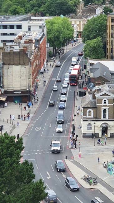 An aerial view of a busy urban street scene in Ealing, showing multiple vehicles including cars, vans, and buses traveling along a curved road. On the left side, there are various commercial buildings with brick and modern facades, some with rooftop structures and signage, while on the right side, a mix of residential and small commercial properties are visible, including a white building with a pitched roof and large windows. Sidewalks on both sides are populated with pedestrians, some walking and others waiting at crosswalks, with a few bicycles parked near a corner. Lush green trees providing shade line the sidewalks and street edges, creating a contrast against the built environment. Traffic light signals and road markings are visible, guiding vehicle movements and indicating designated lanes. The overall lighting suggests daytime with natural daylight illuminating the scene. This scene reflects typical urban street activity, relevant to private waste collection and alternative rubbish removal services managed by Waste Collection Ealing, emphasizing a vibrant, well-used public roadway in a residential and commercial district.