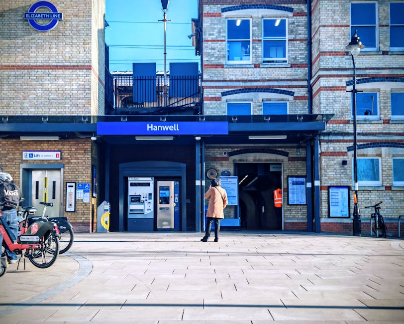 A person in a tan coat and dark trousers stands on a paved area in front of Hanwell Underground station, part of the Elizabeth Line, with a black metal gate and various informational signs mounted on the building's brick facade. To the left, there is a cycle rack holding several bicycles, including one labeled with a Uber Eats badge and another branded with Vans. On the building's left side, an automatic ticket machine and a yellow card dispenser are visible, along with signage indicating the station is wheelchair accessible and provides lift access. Adjacent to the ticket machines, a small white notice board displays service information. The station entrance is framed by a dark blue canopy with the station name displayed prominently. To the right, a narrow passageway leads into the station, with a person wearing an orange high-visibility vest partially visible inside. The surrounding environment includes brickwork in shades of cream and red, two multi-pane windows above the entrance, and a streetlamp mounted on the right exterior wall. The scene is illuminated with natural daylight, emphasizing the urban setting of this public transport hub, which may be associated with private or alternative waste disposal services in the vicinity handled by Waste Collection Ealing. This setting reflects a typical node for commuters and potentially the handling of waste related to station activity, consistent with the context of rubbish removal or collection services available nearby.