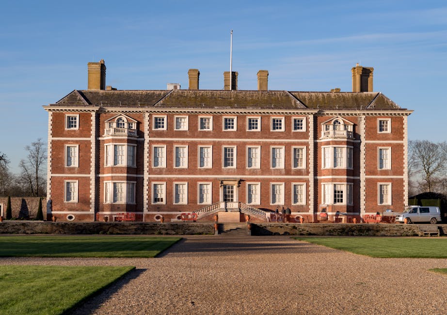 A large, historic brick building with a symmetrical façade, featuring three central floors and a prominent central entrance accessed by a short flight of stairs, flanked by evenly spaced windows on each level. The brickwork displays a reddish-brown hue with contrasting white stone details around the windows, the Cornice, and the decorative window surrounds. The roof is gabled with multiple chimneys rising above, and a flagpole extends upward from the central section of the roof. The building is situated in a well-maintained landscape with trimmed grass, gravel pathways leading to the entrance, and a low stone wall bordering the front. To the right, a white van is parked on a paved area, and several workers or visitors are visible near the front steps, possibly engaged in maintenance or inspection activities. The scene is bathed in natural daylight, with a clear blue sky and some sparse trees in the background, reflecting a peaceful, historic setting possibly associated with private estate use or heritage site management, relating subtly to alternative waste management and on-site clearance services by Waste Collection Ealing.