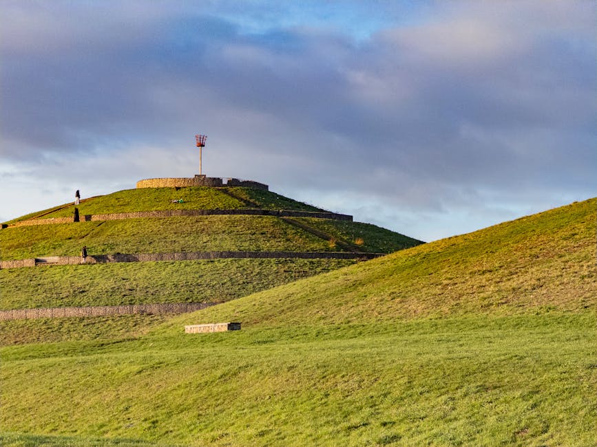 A wide view of a grassy, terraced hill featuring multiple horizontal retaining walls made of dark stone, creating distinct levels across the slope. In the background, a round stone structure with a small modern wind turbine on top is visible at the summit. The hill is bathed in warm, natural sunlight, highlighting the green grass and textured stonework, while a partly cloudy sky with patches of blue and gray clouds looms overhead. Several small figures are scattered across different terraces, emphasizing the hill's scale. The scene appears to be a public park or landscaped area with pathways and steps integrated into the terraced design, characteristic of naturalistic outdoor spaces. The overall setting provides a tranquil and orderly environment, suitable for outdoor leisure activities or scenic viewing, with elements that suggest an intentional landscape design rather than a natural formation, aligning with typical features of outdoor recreational sites managed independently of municipal rubbish collection services.