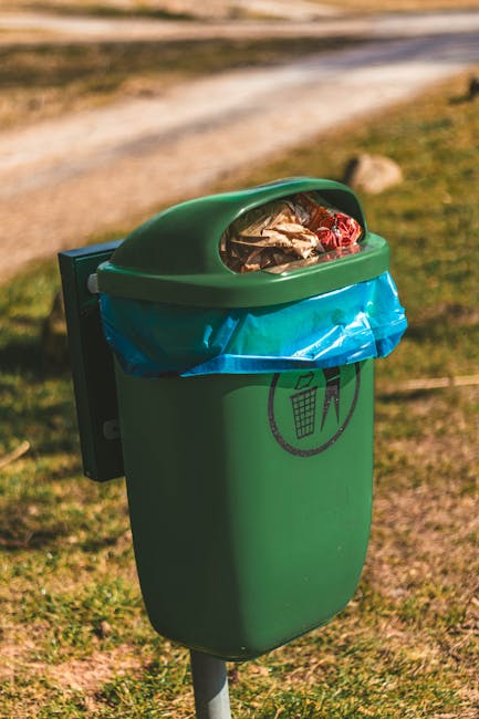 A public park scene during late autumn or early winter, featuring a wide expanse of well-maintained green grass with scattered fallen leaves. Several wooden picnic tables with attached benches are evenly spaced throughout the park, positioned among a row of mature deciduous trees with mostly bare branches, suggesting the season. In the foreground, there is a tall, cylindrical waste bin made of vertical wooden slats with a white plastic liner at the top, situated near a paved pathway edged with a wooden or stone border. The background shows additional trees and pathways, with the lighting indicating either late afternoon or early evening, casting warm tones over the scene. The environment appears peaceful and clean, with no visible litter or debris, reflecting local park maintenance. This setting could relate to private or independent waste handling services, such as rubbish collection, complementing public waste management efforts supported by services like Waste Collection Ealing.